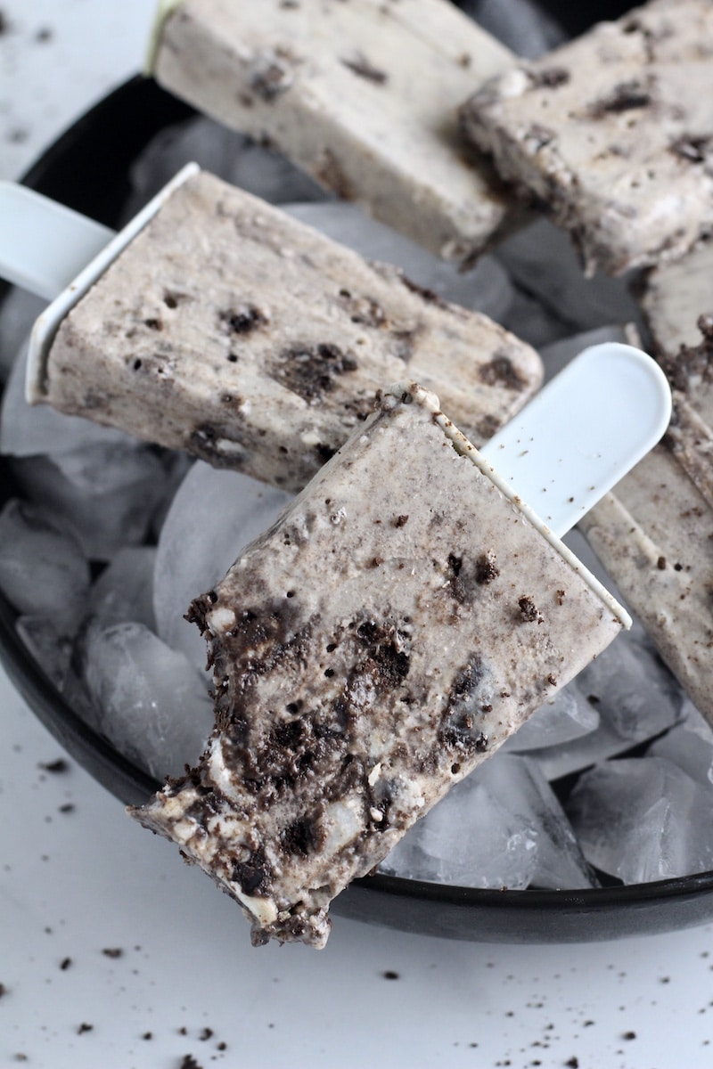 A black plate with ice and cookies and cream popsicles with one bite taken out of the front popsicle.