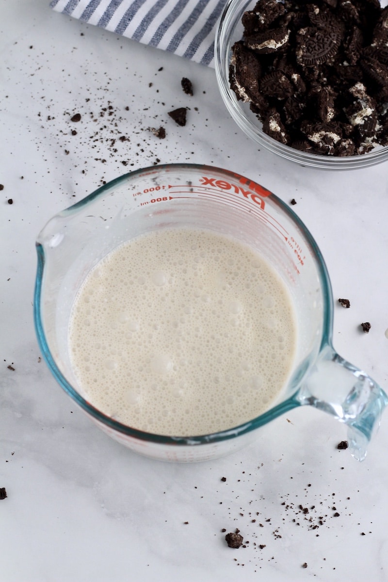 A liquid measuring cup with oat milk and coconut cream and a bowl of Oreo crumbs in the back.