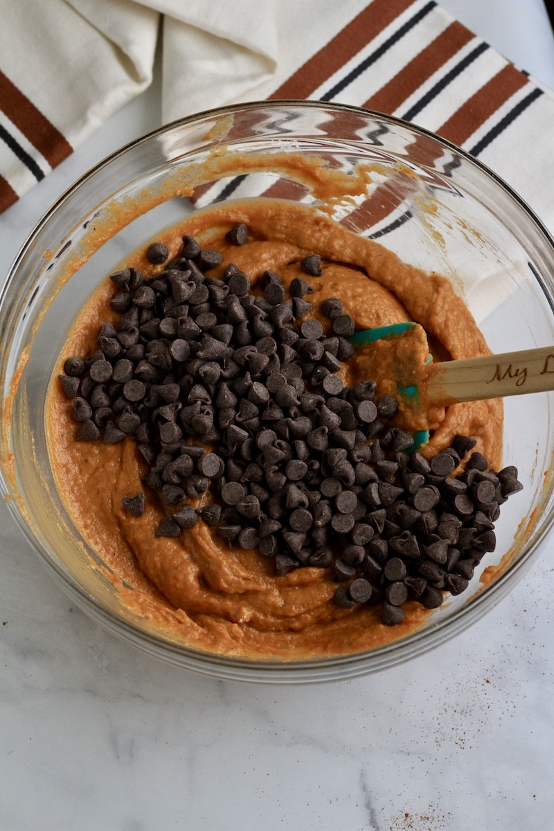 A glass bowl with pumpkin muffin batter topped with chocolate chips before stirring.