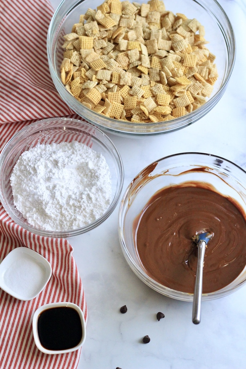 Three glass bowls one with Chex, one with powdered sugar, and one with the cookie butter chocolate mixture with small bowls of vanilla and salt to the side.