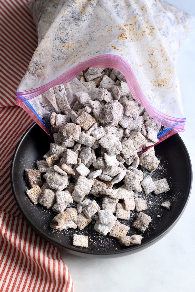 A gallon ziplock pouring cookie butter muddy buddies into a black bowl with a red and white striped towel to the left.