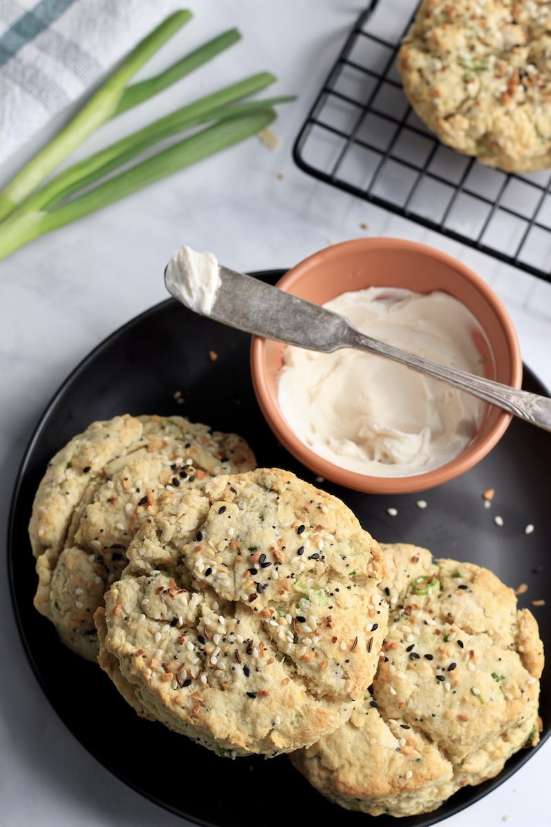 A black plate with three everything bagel biscuits and an orange bowl with dairy-free cream cheese with a knife in front of a cooling rack with more biscuits.