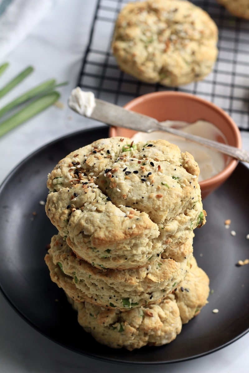 A stack of three biscuits on a black plate with a bowl of dairy-free cream cheese and a cooling rack in the back.