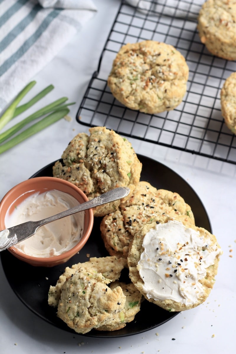 A plate with everything bagel scallion biscuits and a small bowl of cream cheese with a cooling rack of biscuits in the back.