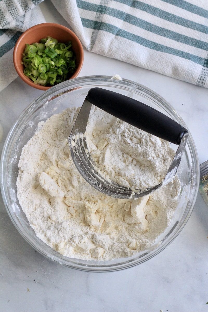 A glass bowl with a pastry blender cutting the vegan butter into the flour and a small bowl of scallions in the back.