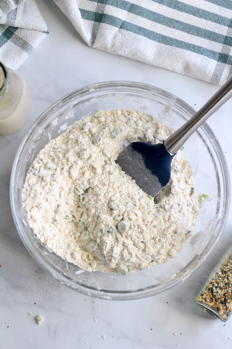 A glass bowl with the dry ingredients mixed together with a blue and silver spatula.