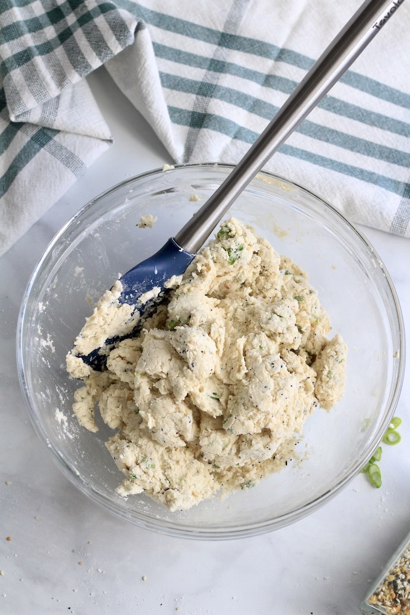 Everything bagel scallion biscuit dough in a glass bowl with a blue and silver spatula.