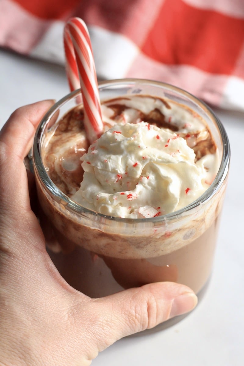 Homemade peppermint hot chocolate in a glass mug with a hand on the left side.