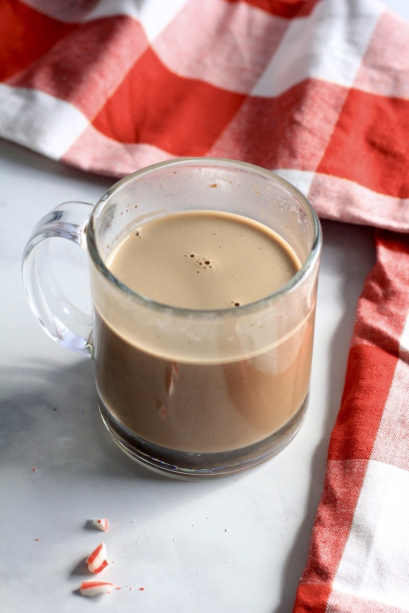 A glass mug with oat milk and melted chocolate chips on a white counter with a red and white buffalo check towel in the back.