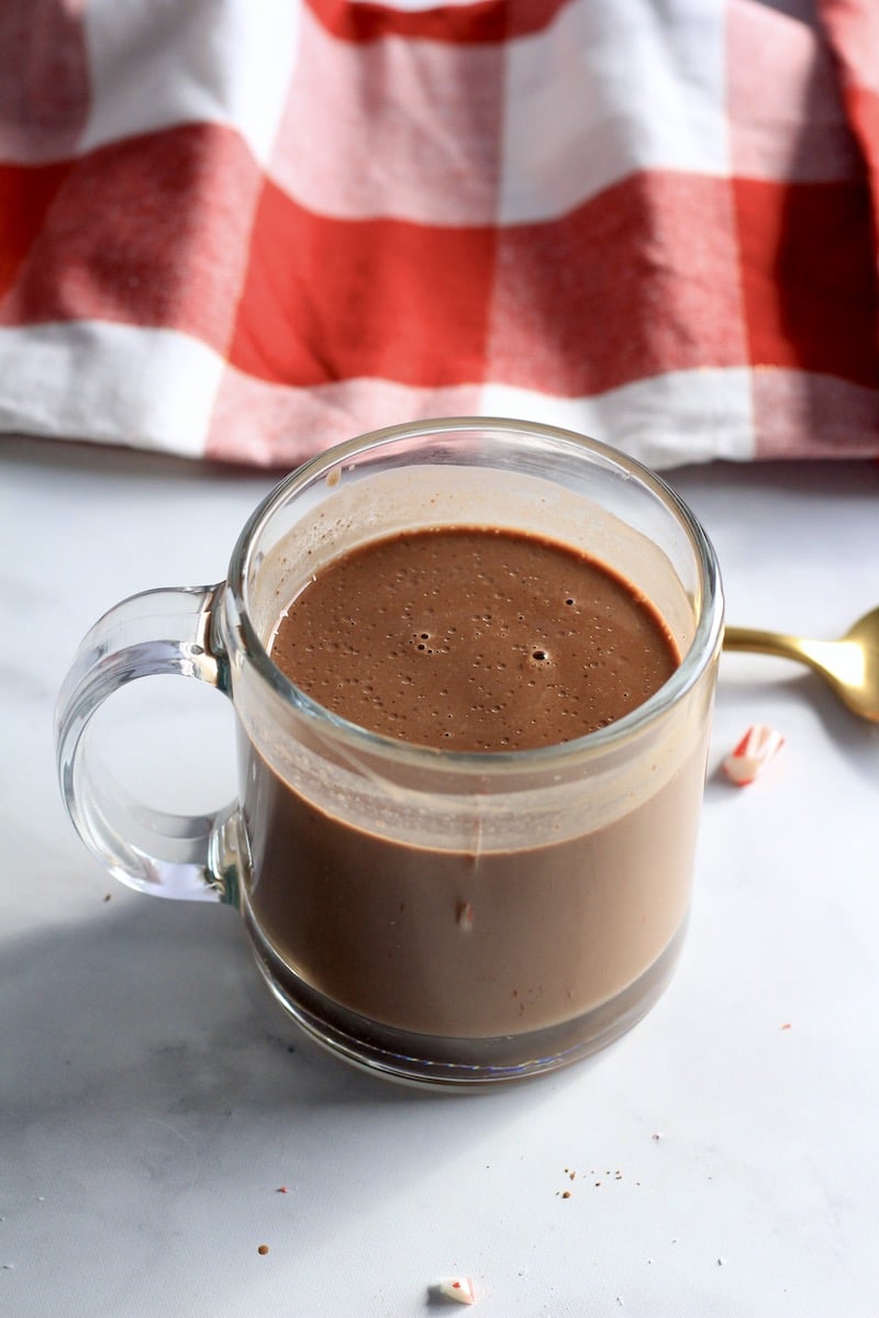 Peppermint hot chocolate in a glass mug on a white counter with a red and white buffalo check towel in the background.