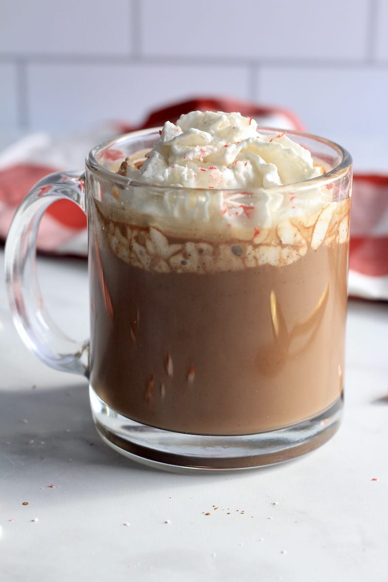 A glass of vegan peppermint hot cocoa on a counter topped with whipped cream and crushed peppermint.