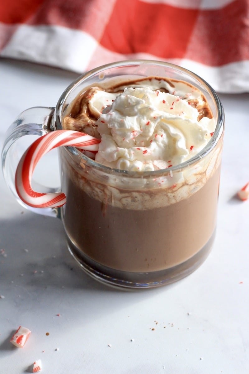 A candy cane sticking out of a glass mug of dairy-free peppermint hot chocolate on a white counter with a red and white towel in the back.
