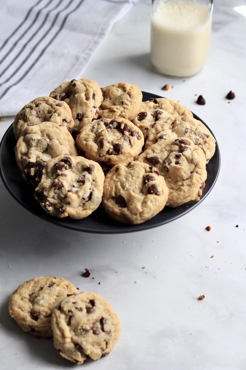 Two chocolate chip cookies on the counter to left left with a plate of cookies in the middle and a bottle of flax milk in the back right.