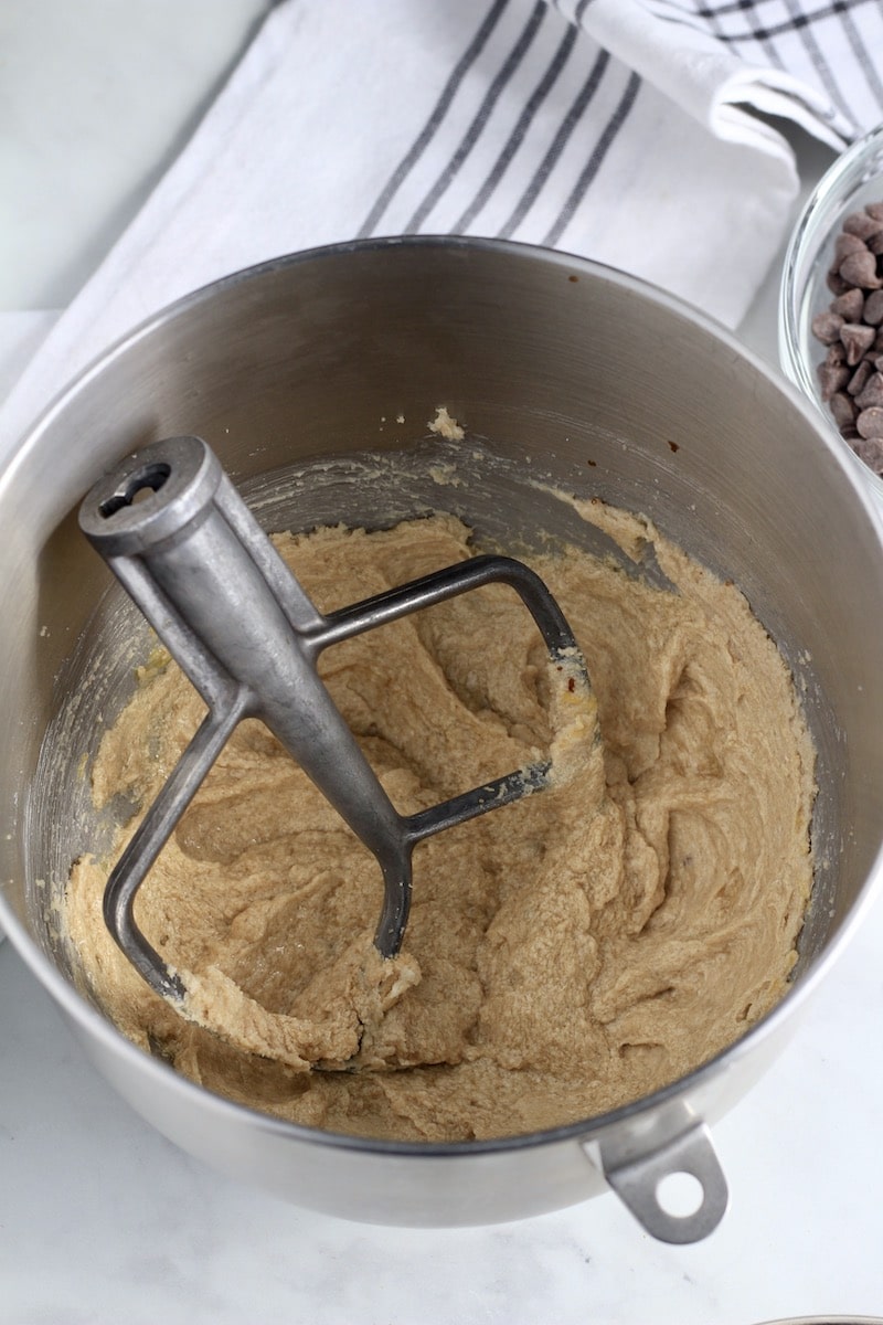 A silver mixing bowl with wet ingredients creamed together and a silver paddle attachment on the left side.