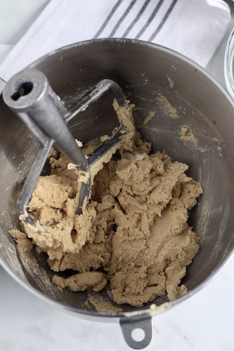 A silver mixing bowl with cookie dough and a paddle attachment on the left side.