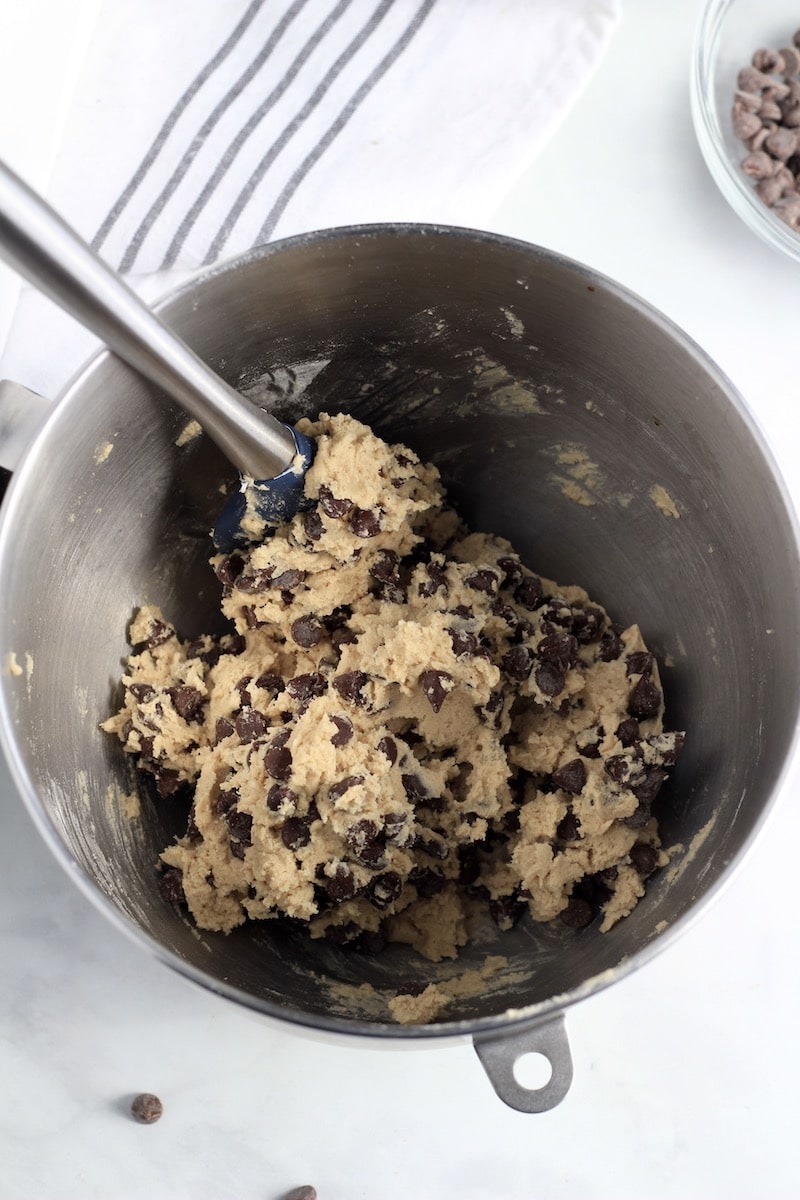 A silver mixing bowl with chocolate chip cookie dough and a spatula in the top left of the bowl.