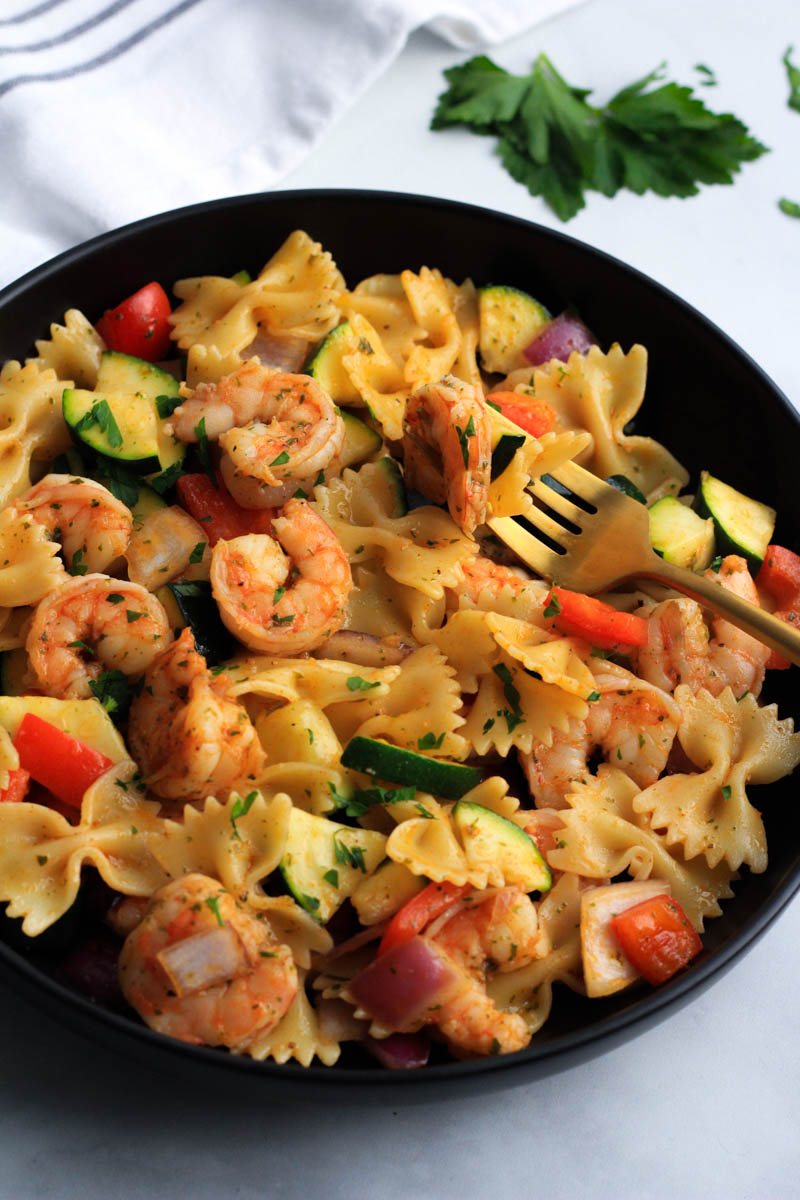 A close up of a black bowl with shrimp and veggie pasta on a gold fork.