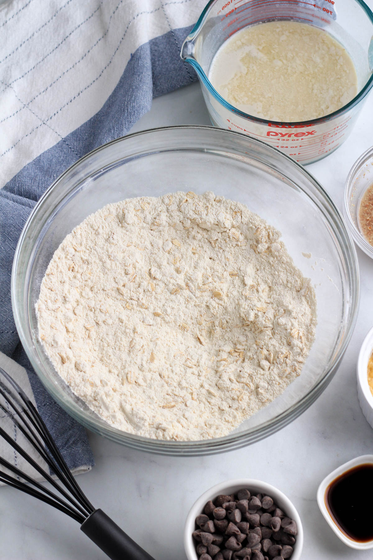 A large glass bowl with the mixed dry ingredients with the remaining ingredients around the outside of the bowl.