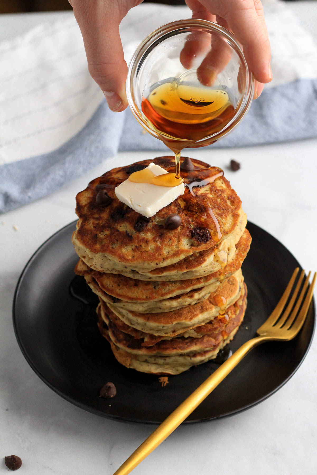 A hand pouring a small glass bowl with maple syrup over a stack of vanilla chocolate chip vegan pancakes on a black plate.