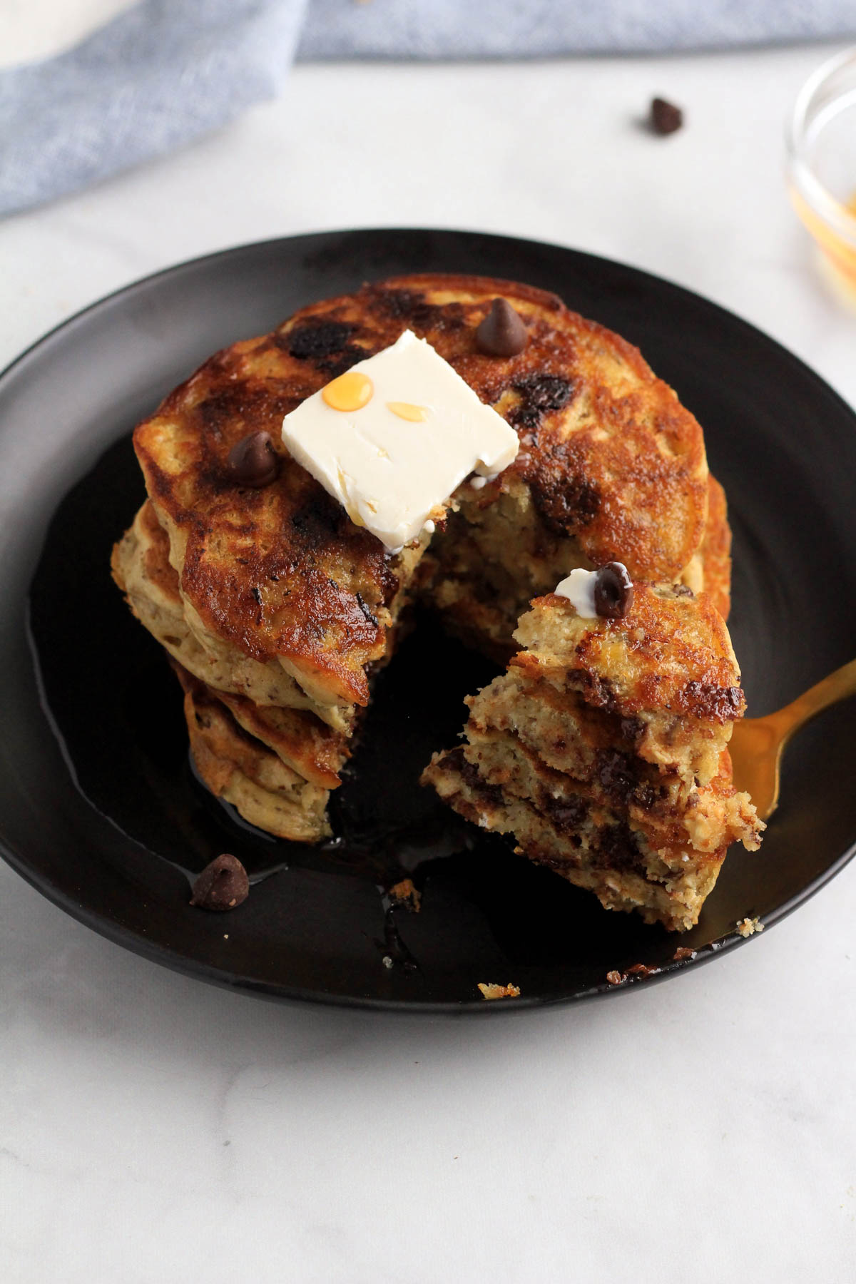 A top down photo of a stack of vegan chocolate chip pancakes on a black plate with a triangle bite on a gold fork on the right.