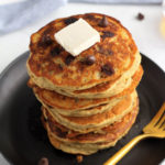 A black plate with a stack of chocolate chip pancakes topped with vegan butter and a gold fork to the right.