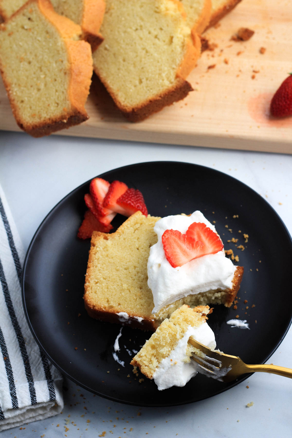 A slice of dairy-free vanilla pound cake on a plate topped with dairy-free whipped topping and sliced strawberries with a fork that has a bite of cake on it.