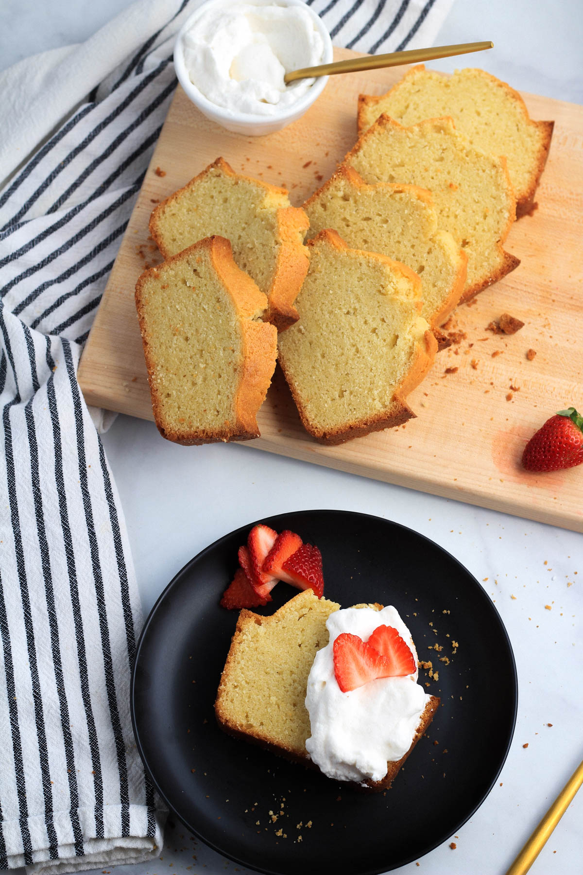 A small plate with a slice of vanilla pound cake topped with dairy-free whipped cream and strawberries with cutting board in the back with more slices of pound cake.
