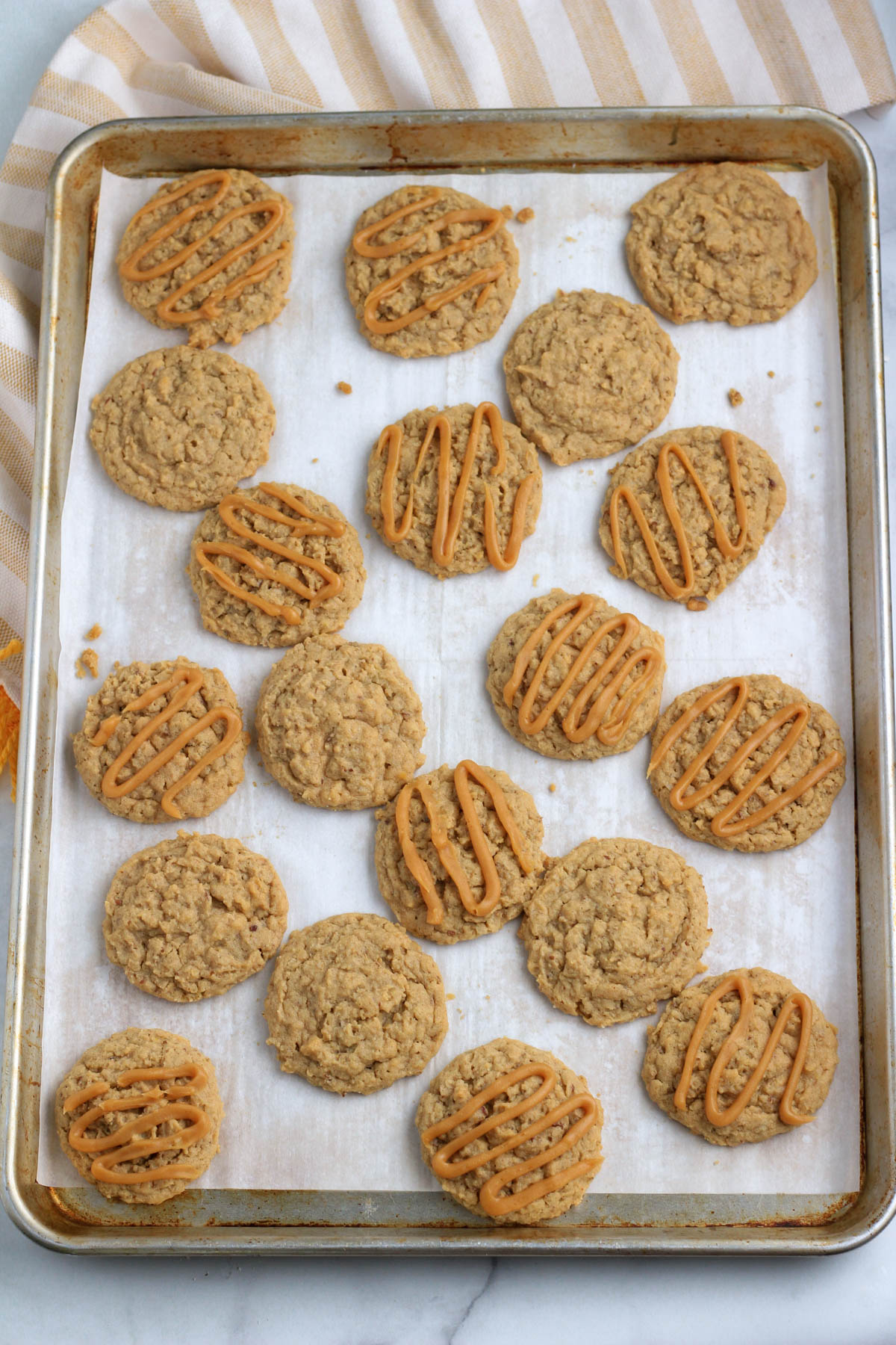 A silver rimmed cookie sheet on a counter with a yellow and white striped towel topped with vegan peanut butter oatmeal cookies.