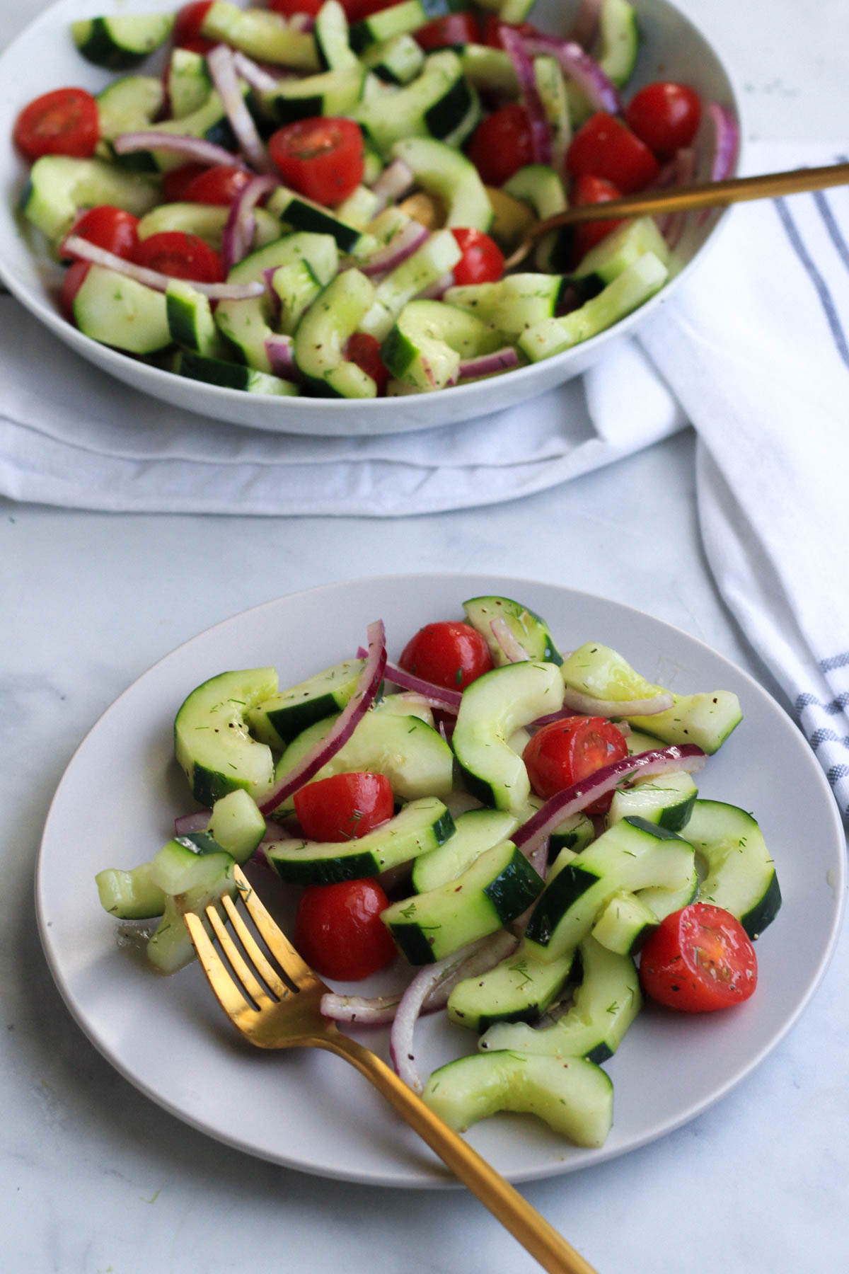 Vegan cucumber and dill salad on a small white plate with a gold fork and a bowl of salad in the back.