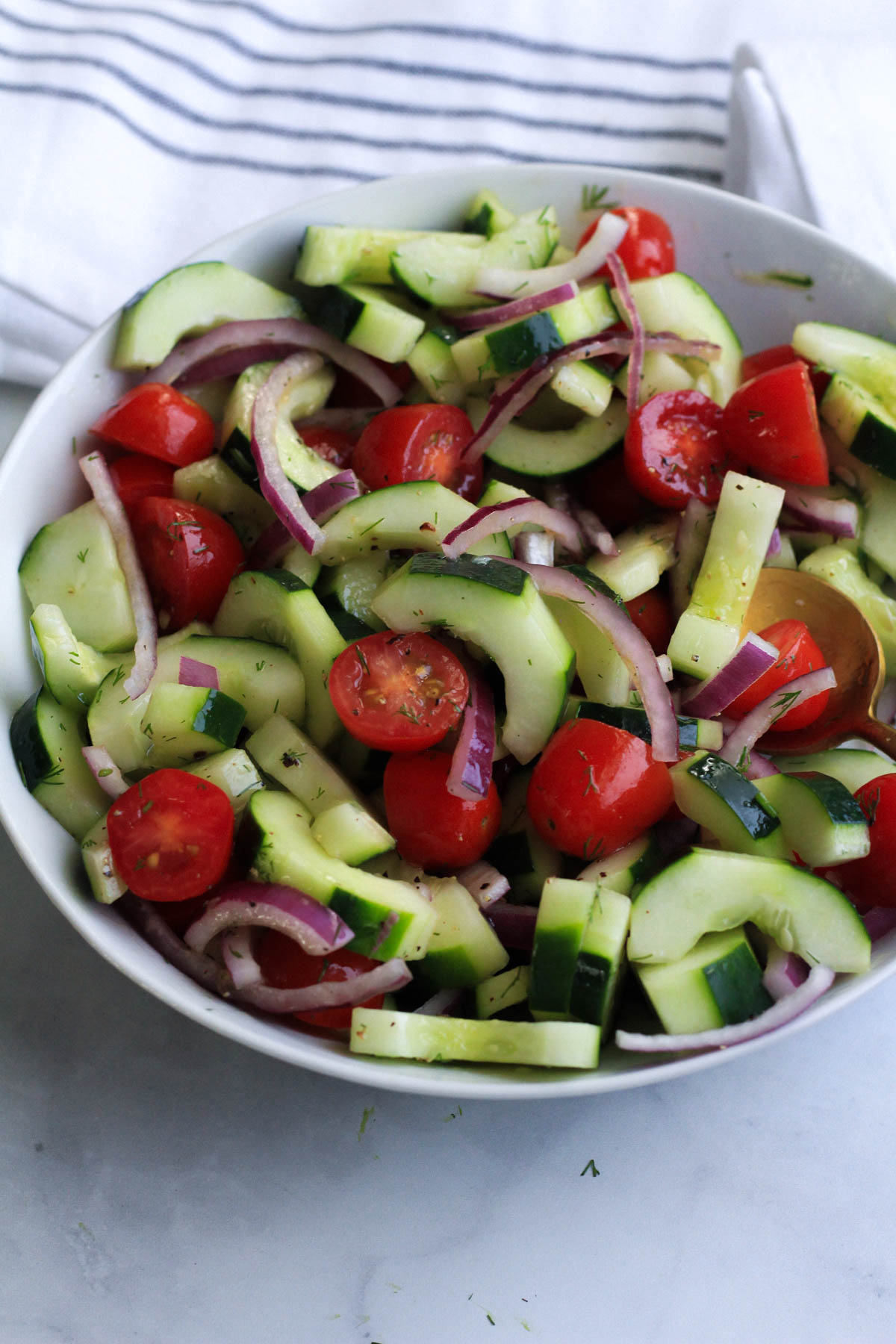 A cucumber and dill salad tossed in a white bowl with a white and blue towel in the background.