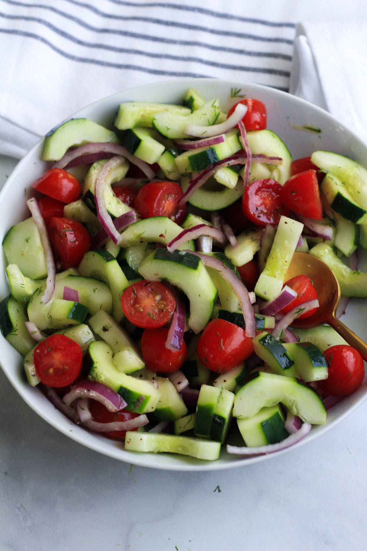 Vegan cucumber and dill salad tossed together in a white bowl with a blue and white striped towel in the back.