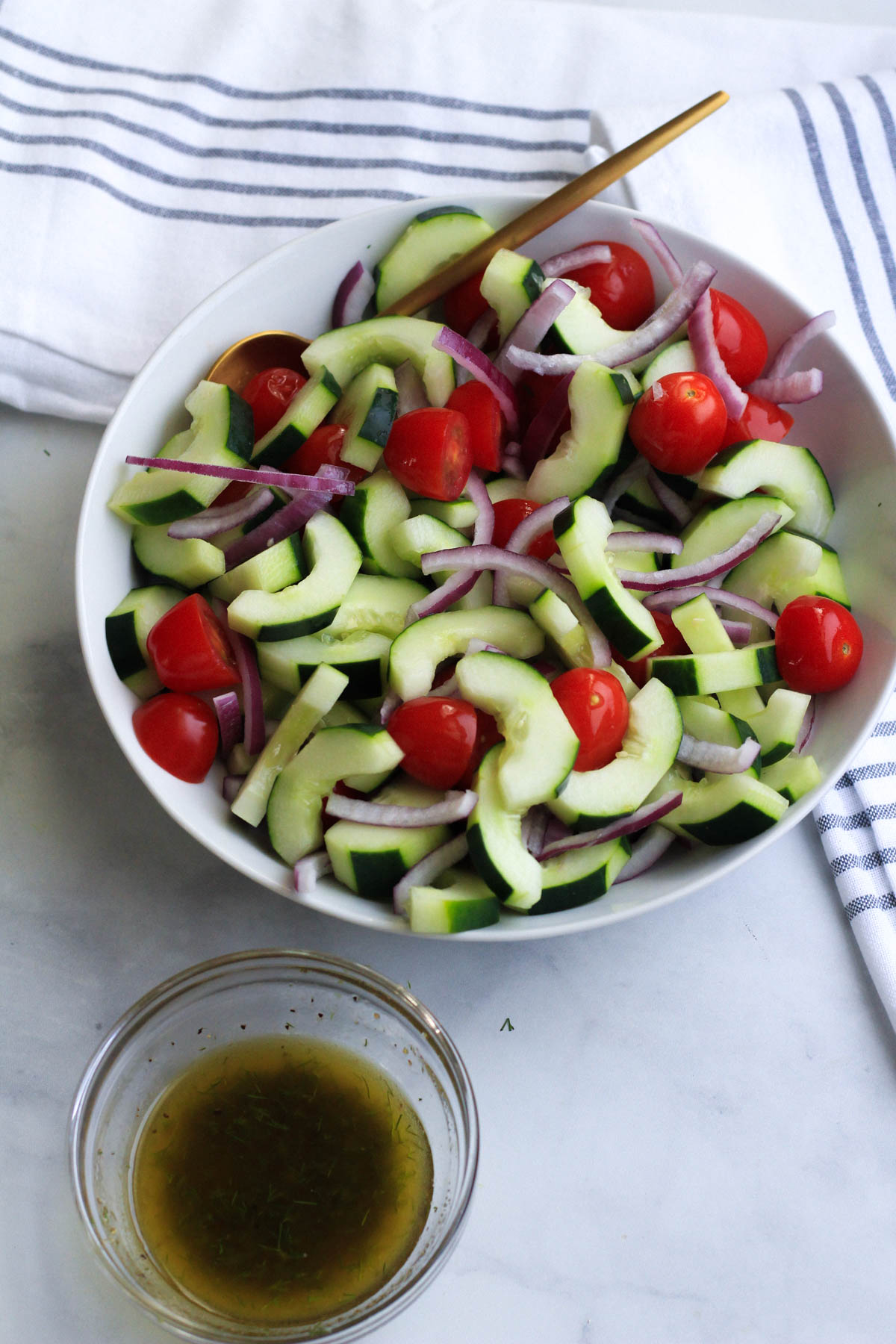 A white bowl with cucumber and dill salad vegetables and a gold spoon in the top with the dressing in the front.