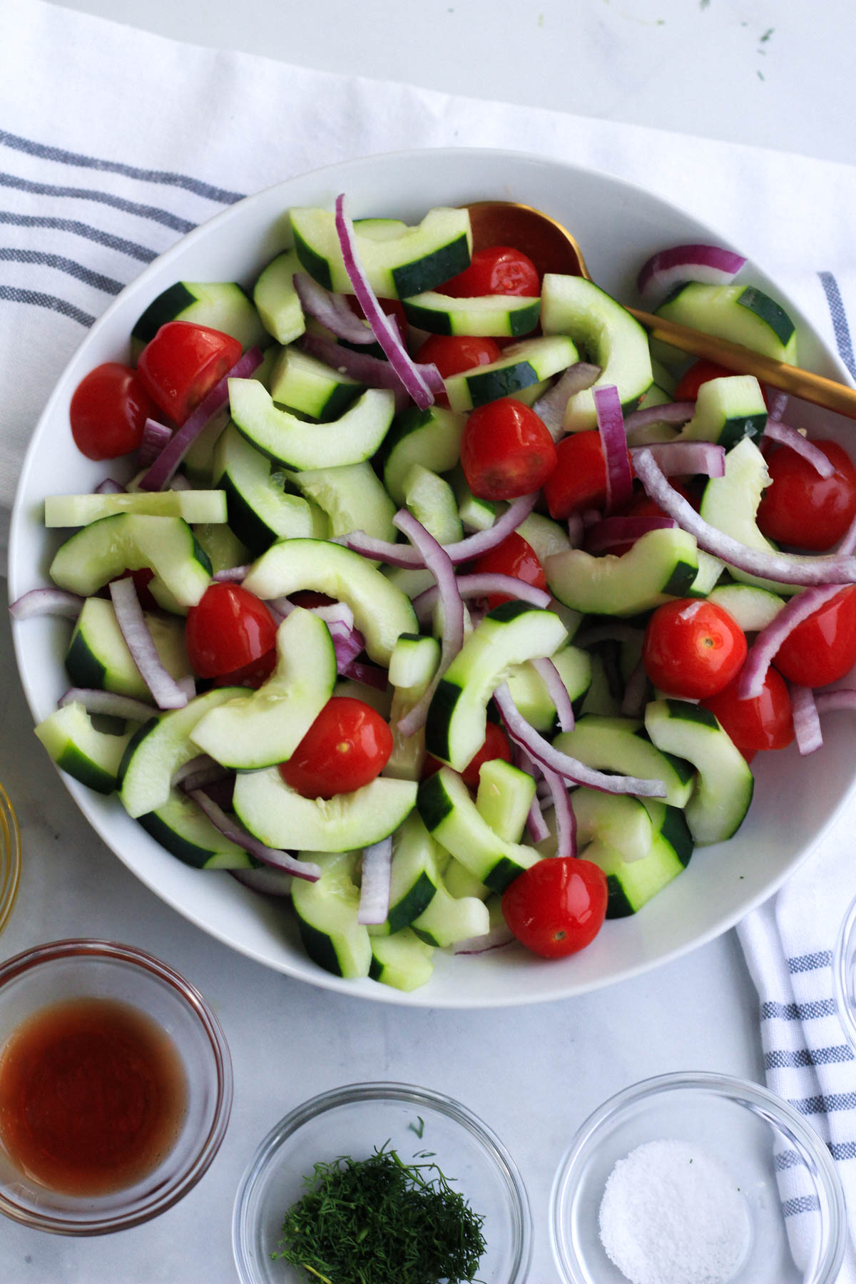 A white bowl with the cucumber salad ingredients mixed together and some of the dressing ingredients in the front in small glass bowls.