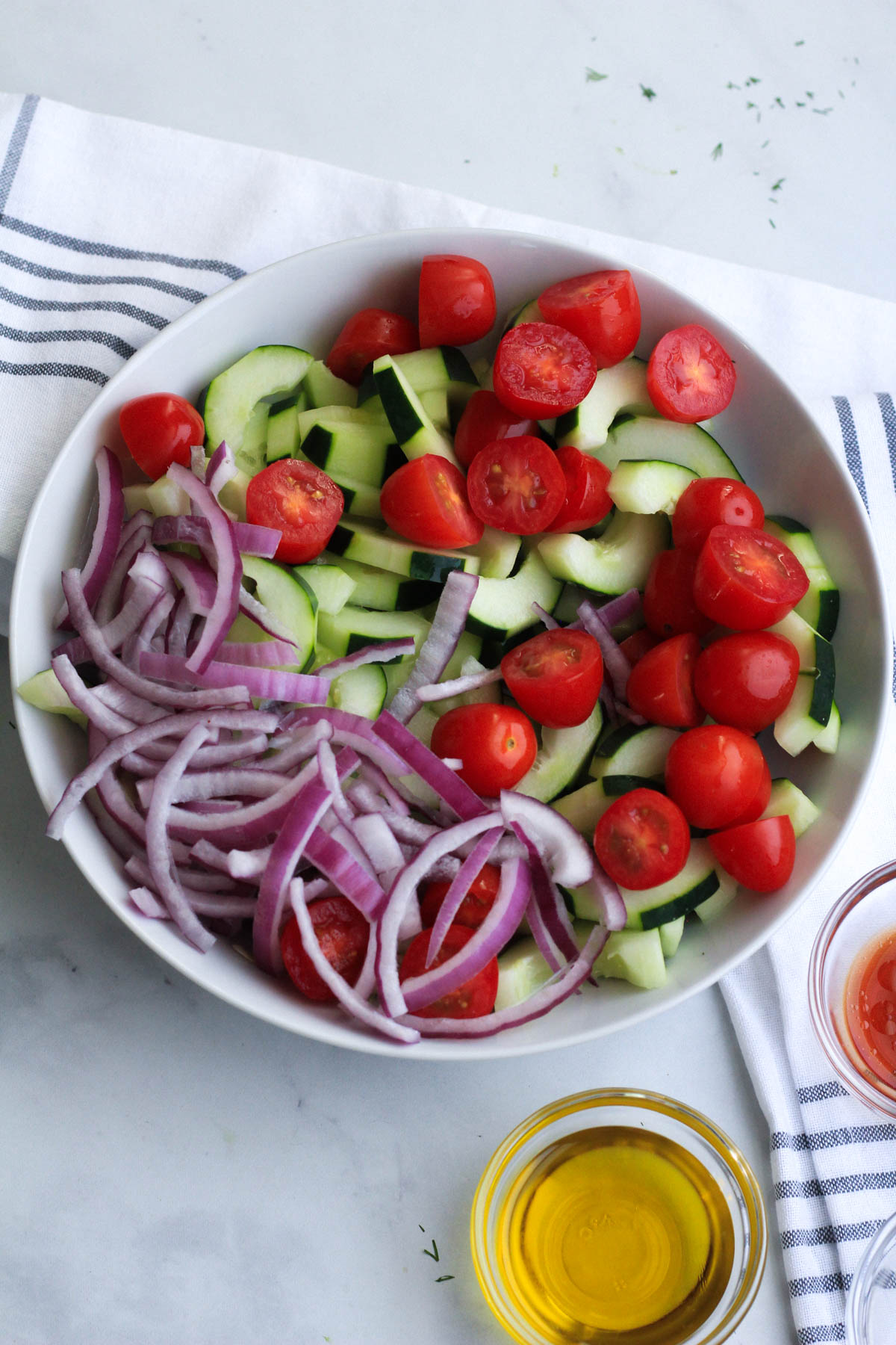 A white bowl with the vegetables for cucumber dill salad and a small bowl of dressing ingredients in the front.