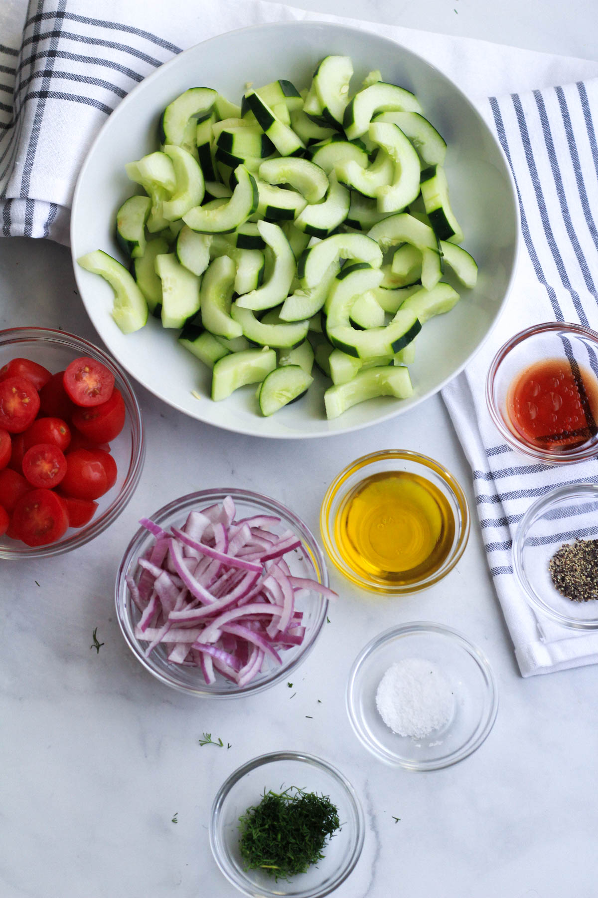 The ingredients for cucumber and dill salad on a white counter with a blue and white towel in the top.