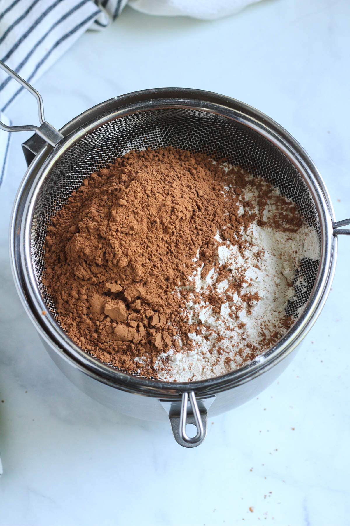 A fine mesh strainer topped with dry ingredients over the silver mixing bowl.