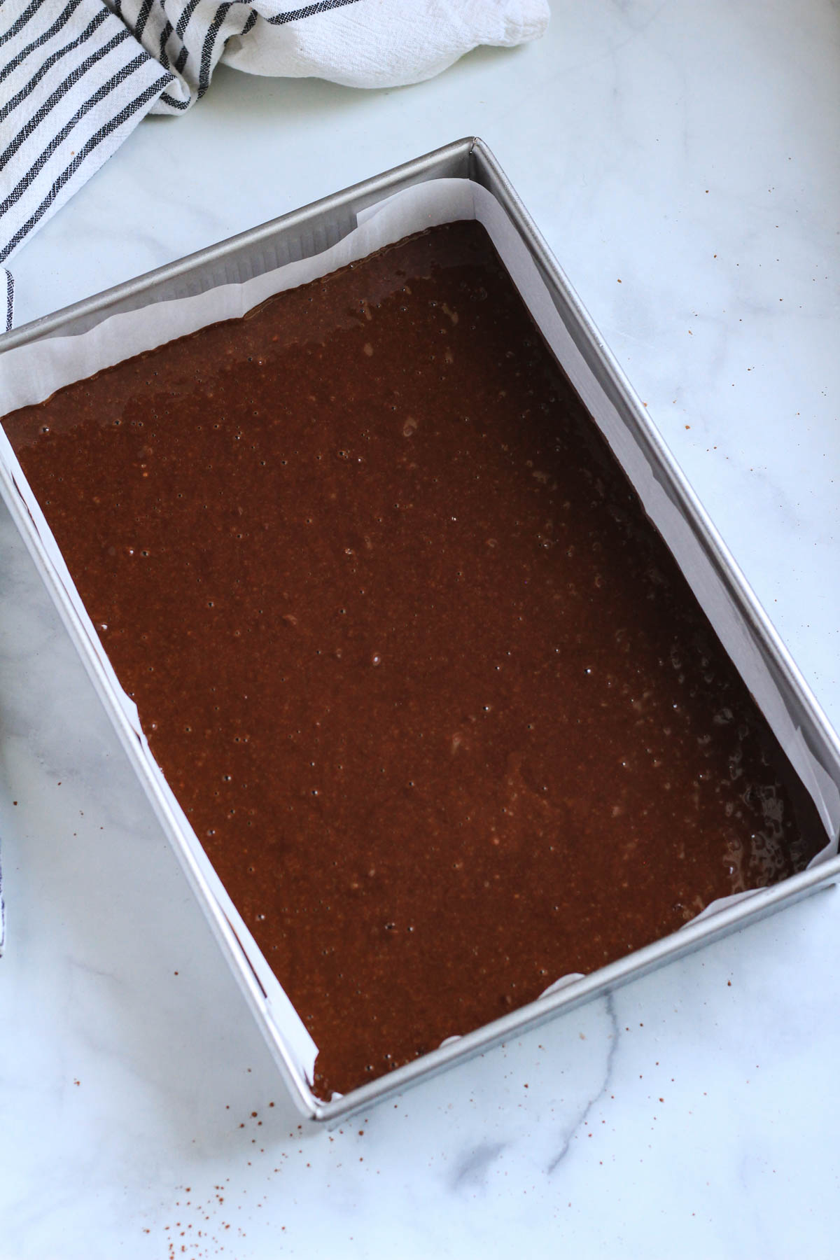 A silver rectangular baking pan lined with parchment paper and filled with dairy-free chocolate cake batter.