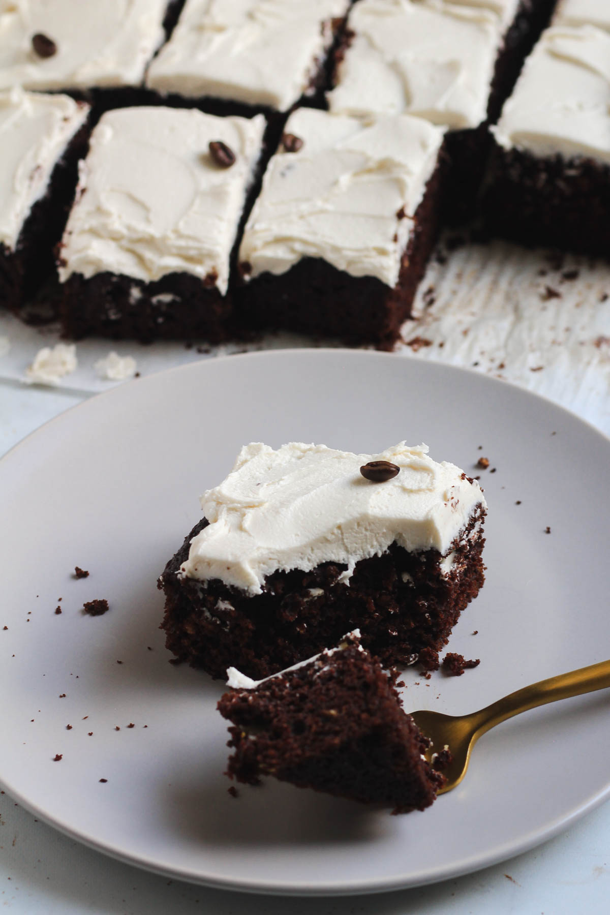 A square of frosted chocolate cake on a white plate with cake slices in the back.