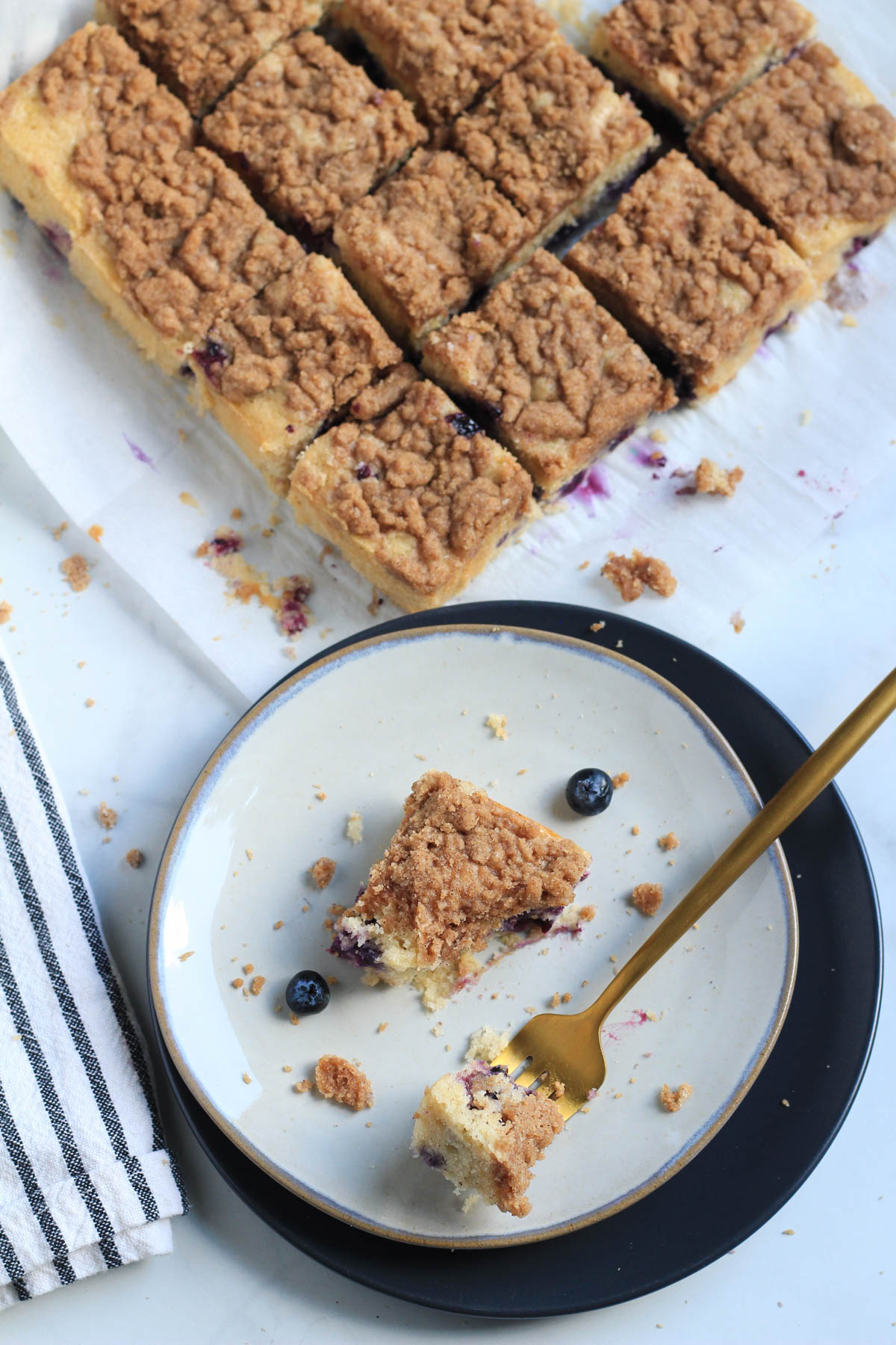 A plate of coffee cake with a bite on a fork in the front and a parchment paper with squares of coffee cake in the back.