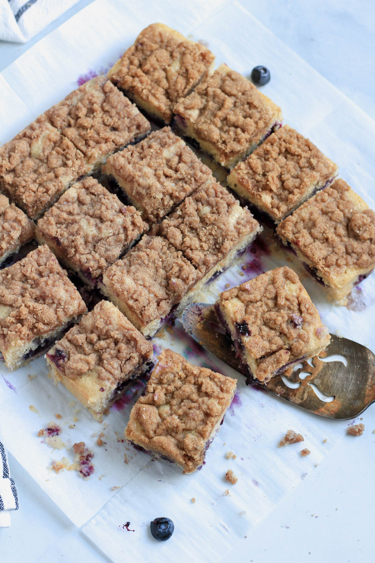 A parchment paper with lemon blueberry coffee cake cut into squares and a silver spatula on the right with a slice of cake ready to serve.