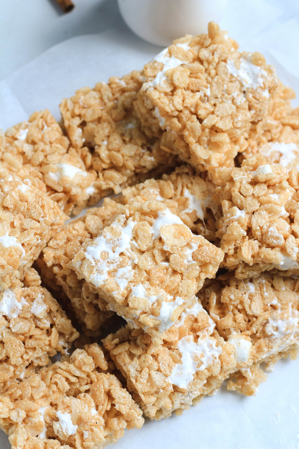 A pile of cinnamon maple Rice Krispie treats on a piece of parchment paper with a small jug of maple syrup in the back.