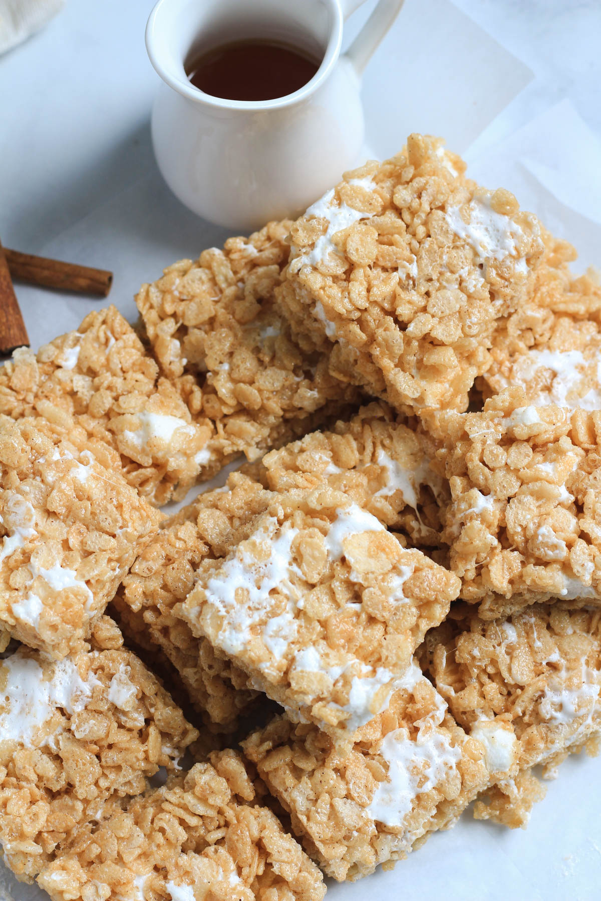 A top down of maple Rice Krispie treats on parchment paper with a jug of maple syrup and some cinnamon sticks in the back.