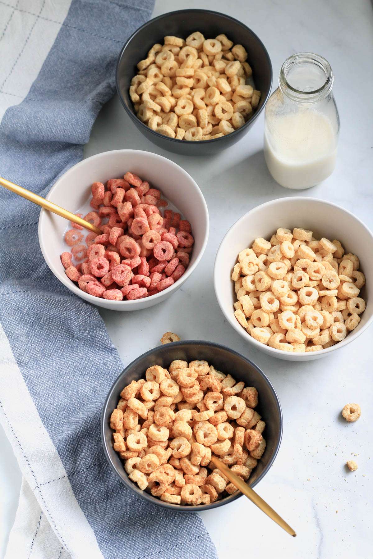 Four bowls of three wishes cereal on a white counter with a blue and white striped towel on the left side and a bottle of non-dairy milk in the top right.