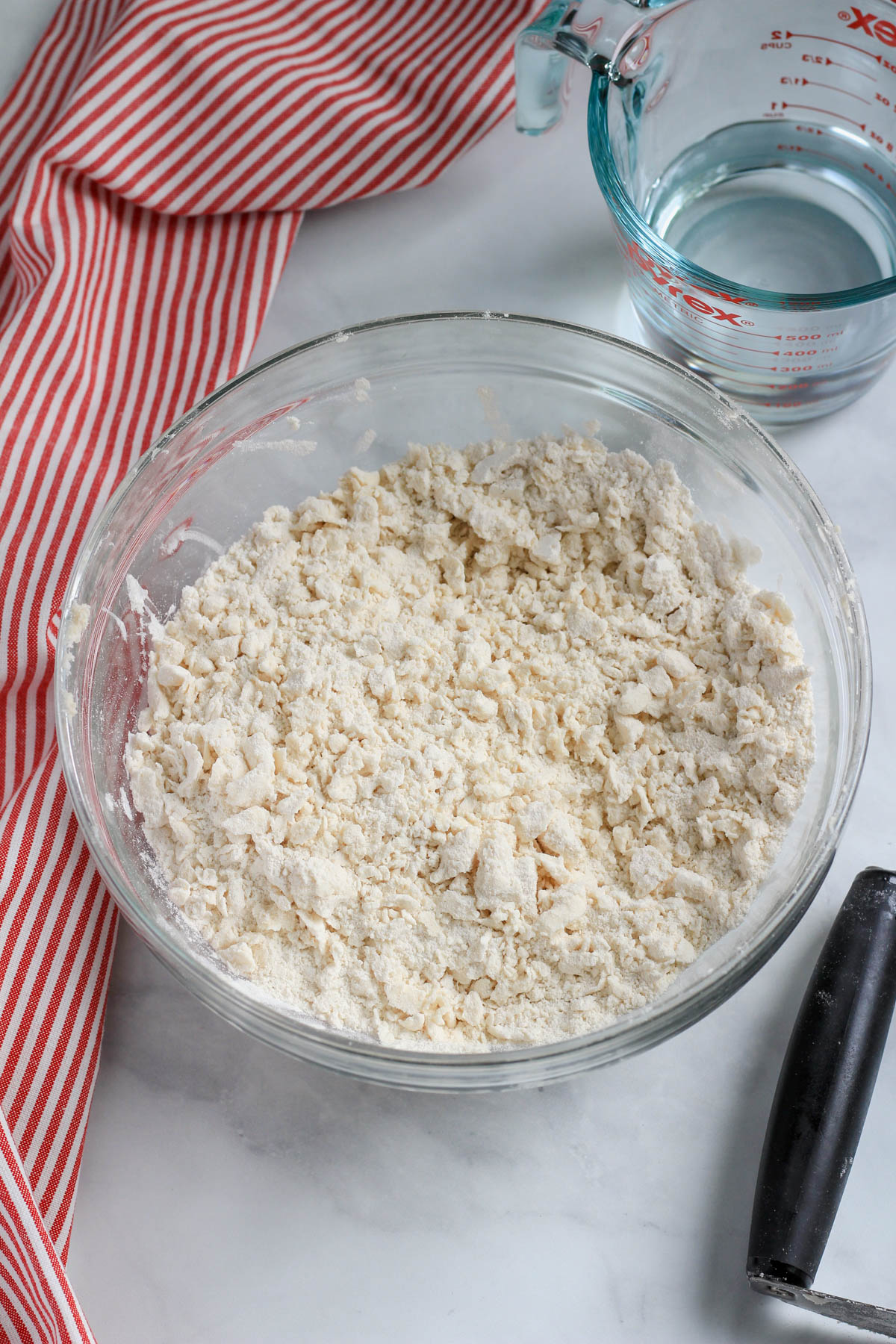 A glass bowl witha flour mixture and vegan butter cut in with a pastry blender in the bottom right and a liquid measuring cup in the top right with a red and white striped dish towel on the left side.