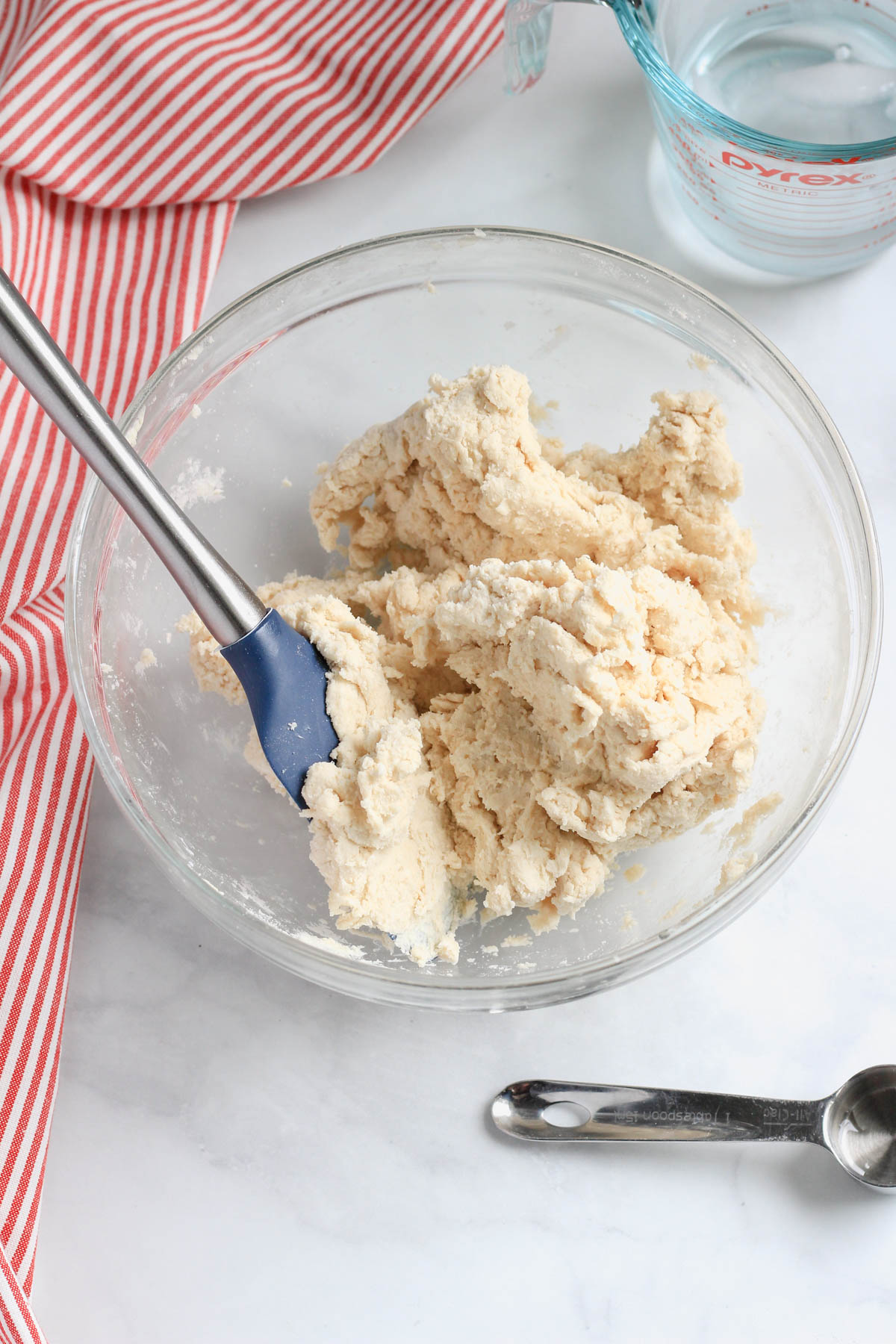 A glass bowl with a silver and blue rubber spatula and a loosely formed dough ball in the bowl with a tablespoon in the bottom right of the photo.