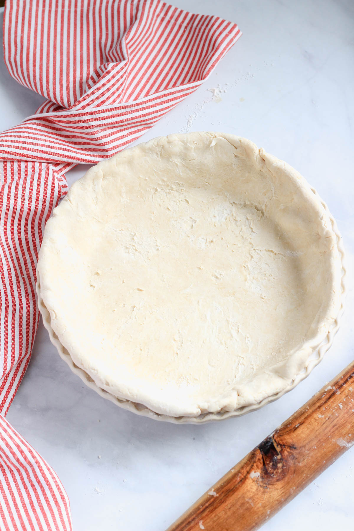 A vegan all butter pie crust in a pie pan with crimped edges and a wooden rolling pin in the bottom right.