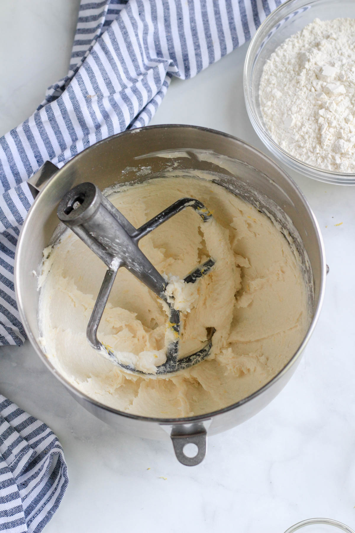 A silver mixing bowl with a paddle attachment with creamed plant-butter and sugars on a white counter.