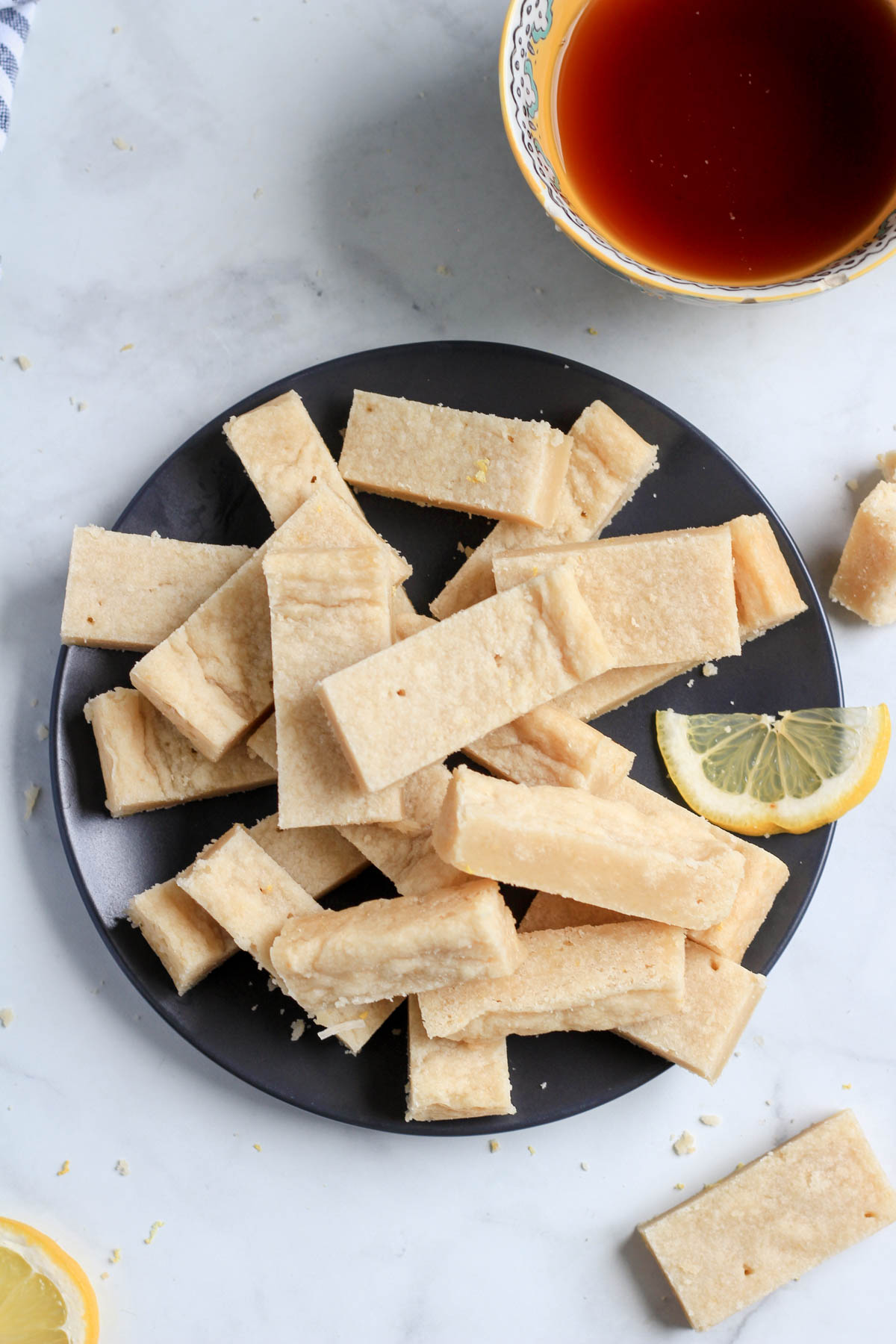 A blue plate of cut bars of lemon shortbread with a small slice of lemon on the right side and a cup of tea in the top right corner.