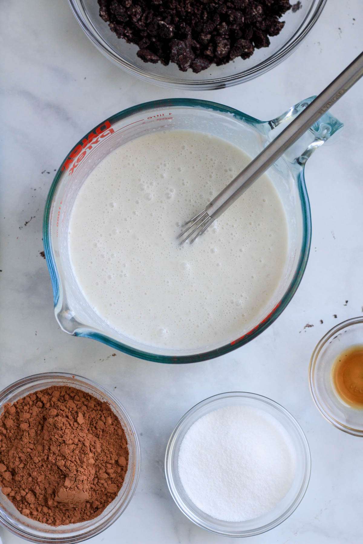 A liquid measuring cup with oat milk and coconut milk mixed together with remaining ingredients in bowls around.