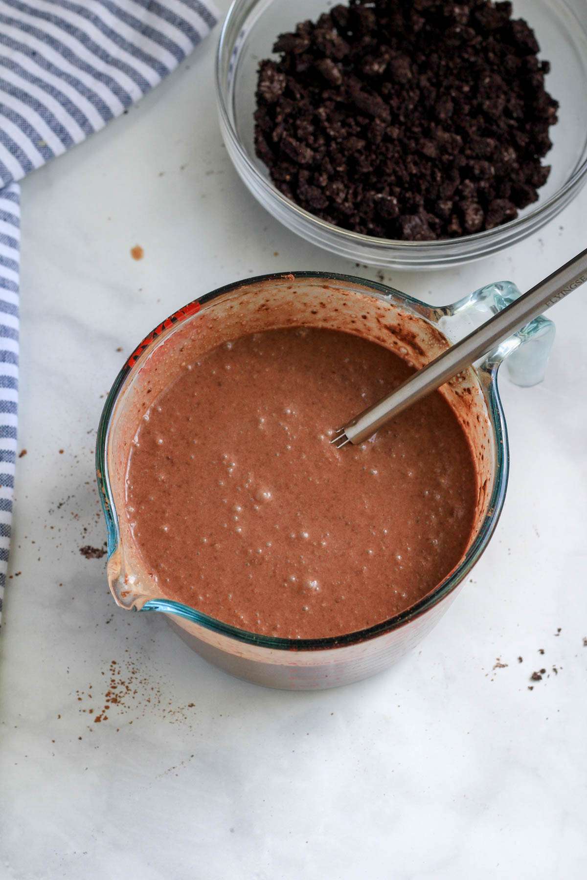 A large liquid measuring cup with a small whisk and vegan fudgesicle filling with a bowl of Oreo crumbs to the top right.